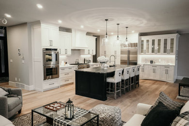 Example of a large transitional l-shaped light wood floor and brown floor open concept kitchen design in Edmonton with a double-bowl sink, shaker cabinets, white cabinets, granite countertops, beige backsplash, cement tile backsplash, stainless steel appliances, an island and black countertops