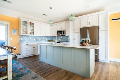Example of a large beach style l-shaped vinyl floor and brown floor eat-in kitchen design in Other with a double-bowl sink, recessed-panel cabinets, white cabinets, quartz countertops, blue backsplash, glass tile backsplash, stainless steel appliances, an island and white countertops