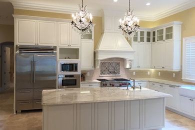 Large transitional l-shaped limestone floor open concept kitchen photo in Miami with a farmhouse sink, raised-panel cabinets, white cabinets, marble countertops, beige backsplash, glass tile backsplash, stainless steel appliances and an island