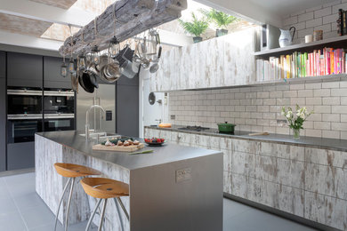 This is an example of a large industrial galley eat-in kitchen in London with an integrated sink, flat-panel cabinets, stainless steel benchtops, white splashback, ceramic splashback, stainless steel appliances, with island and grey floor.