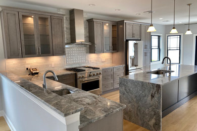 Photo of a mid-sized beach style l-shaped open plan kitchen in New York with an undermount sink, shaker cabinets, grey cabinets, granite benchtops, grey splashback, glass tile splashback, stainless steel appliances, light hardwood floors, with island, white floor and grey benchtop.