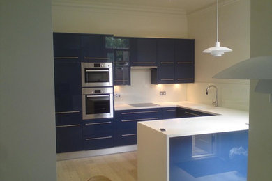 Photo of a large victorian u-shaped kitchen in London with an integrated sink, flat-panel cabinets, blue cabinets, composite countertops, white splashback, glass sheet splashback, stainless steel appliances, light hardwood flooring and an island.