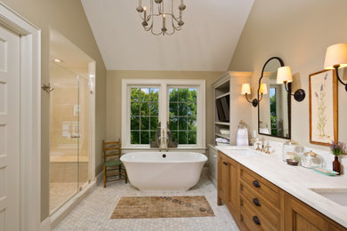 Photo of a medium sized classic ensuite bathroom in New York with medium wood cabinets, a freestanding bath, beige walls, ceramic flooring, a submerged sink, marble worktops, grey floors, a hinged door, white worktops, a shower bench, double sinks, a freestanding vanity unit and recessed-panel cabinets.