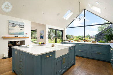 Design ideas for an expansive contemporary u-shaped kitchen/diner in Berkshire with a belfast sink, blue cabinets, medium hardwood flooring, an island, brown floors, white worktops, a vaulted ceiling and a chimney breast.