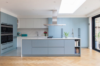 Design ideas for a medium sized contemporary l-shaped open plan kitchen in London with a built-in sink, flat-panel cabinets, blue cabinets, composite countertops, white splashback, marble splashback, black appliances, ceramic flooring, an island, multi-coloured floors and white worktops.