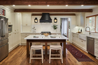 Large elegant u-shaped medium tone wood floor, brown floor and exposed beam enclosed kitchen photo in Portland with a farmhouse sink, shaker cabinets, beige cabinets, quartz countertops, white backsplash, wood backsplash, stainless steel appliances, an island and white countertops