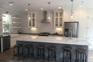 Inspiration for a mid-sized transitional l-shaped medium tone wood floor eat-in kitchen remodel in Orange County with a farmhouse sink, shaker cabinets, white cabinets, marble countertops, white backsplash, brick backsplash, stainless steel appliances and an island