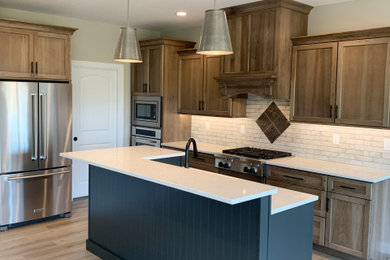 Mid-sized elegant single-wall medium tone wood floor and brown floor eat-in kitchen photo in Other with a farmhouse sink, recessed-panel cabinets, brown cabinets, quartz countertops, white backsplash, stone tile backsplash, stainless steel appliances, an island and white countertops