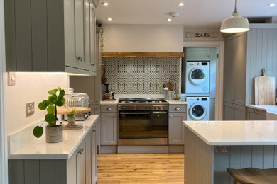 Photo of a medium sized country kitchen/diner in Wiltshire with a belfast sink, shaker cabinets, grey cabinets, quartz worktops, multi-coloured splashback, ceramic splashback, stainless steel appliances, medium hardwood flooring, a breakfast bar, white worktops and feature lighting.