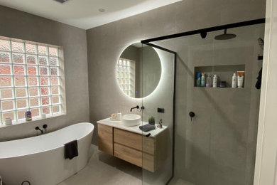 This is an example of a large contemporary kids bathroom in Other with beaded inset cabinets, medium wood cabinets, a freestanding tub, a corner shower, a wall-mount toilet, beige tile, porcelain tile, beige walls, porcelain floors, a vessel sink, solid surface benchtops, beige floor, a hinged shower door, white benchtops, a niche, a single vanity and a floating vanity.