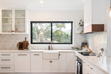 Design ideas for a large country l-shaped kitchen pantry in Melbourne with a farmhouse sink, shaker cabinets, white cabinets, quartz benchtops, beige splashback, porcelain splashback, black appliances, dark hardwood floors, with island, brown floor and white benchtop.