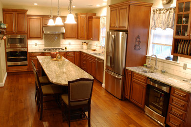 Large elegant l-shaped medium tone wood floor and brown floor eat-in kitchen photo in DC Metro with an undermount sink, raised-panel cabinets, brown cabinets, granite countertops, white backsplash, ceramic backsplash, stainless steel appliances and an island