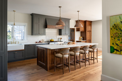 Example of a large transitional l-shaped medium tone wood floor open concept kitchen design in Burlington with a farmhouse sink, shaker cabinets, quartzite countertops, white backsplash, subway tile backsplash, paneled appliances, an island and white countertops