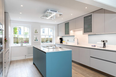 Photo of a mid-sized modern galley open plan kitchen in London with an undermount sink, flat-panel cabinets, grey cabinets, quartzite benchtops, white splashback, stone slab splashback, black appliances, light hardwood floors, with island, brown floor and white benchtop.