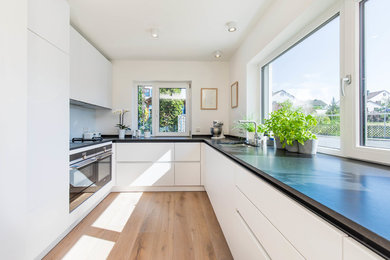 This is an example of a mid-sized modern u-shaped kitchen in Nuremberg with a drop-in sink, flat-panel cabinets, white cabinets, blue splashback, glass sheet splashback, black appliances, light hardwood floors, no island, beige floor and black benchtop.