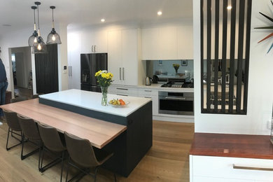 Photo of a large transitional l-shaped open plan kitchen in Melbourne with a double-bowl sink, flat-panel cabinets, white cabinets, quartz benchtops, metallic splashback, mirror splashback, stainless steel appliances, medium hardwood floors, with island, brown floor and white benchtop.
