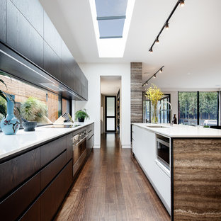 Inspiration pour une grande arrière-cuisine parallèle design avec un évier 2 bacs, un placard à porte plane, des portes de placard en bois sombre, une crédence en feuille de verre, un électroménager en acier inoxydable, un sol en bois foncé, un îlot central, un sol marron et un plan de travail blanc.