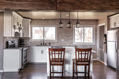 Mid-sized mountain style u-shaped laminate floor and brown floor open concept kitchen photo with an undermount sink, raised-panel cabinets, white cabinets, quartzite countertops, white backsplash, brick backsplash, stainless steel appliances, an island and gray countertops