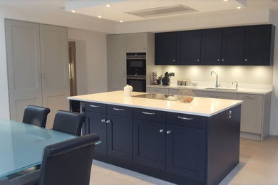 This is an example of a mid-sized traditional l-shaped eat-in kitchen in Surrey with a double-bowl sink, shaker cabinets, blue cabinets, quartzite benchtops, white splashback, glass sheet splashback, black appliances, porcelain floors, with island, grey floor and white benchtop.