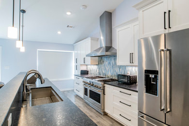 Example of a mid-sized transitional galley porcelain tile, brown floor and vaulted ceiling open concept kitchen design in Other with a single-bowl sink, shaker cabinets, white cabinets, granite countertops, blue backsplash, glass tile backsplash, stainless steel appliances, an island and black countertops