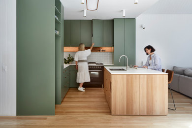 Large contemporary l-shaped open plan kitchen in Melbourne with an undermount sink, flat-panel cabinets, green cabinets, solid surface benchtops, grey splashback, porcelain splashback, black appliances, light hardwood floors, with island, brown floor and white benchtop.