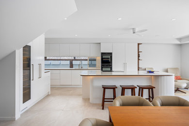 Photo of a large modern galley kitchen pantry in Gold Coast - Tweed with an undermount sink, flat-panel cabinets, white cabinets, quartz benchtops, window splashback, white appliances, porcelain floors, with island, beige floor and white benchtop.