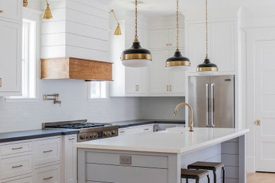 Design ideas for a country l-shaped kitchen in Houston with a farmhouse sink, shaker cabinets, white cabinets, white splashback, subway tile splashback, stainless steel appliances, medium hardwood floors, with island, brown floor and black benchtop.
