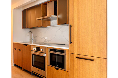 Photo of a mid-sized contemporary single-wall separate kitchen in New York with an undermount sink, glass-front cabinets, medium wood cabinets, quartz benchtops, white splashback, stone slab splashback, stainless steel appliances, light hardwood floors, no island, beige floor and white benchtop.