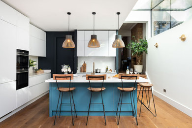 Design ideas for a contemporary l-shaped kitchen in London with an undermount sink, flat-panel cabinets, white cabinets, black appliances, medium hardwood floors, with island, brown floor and white benchtop.