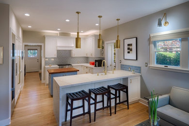 Photo of a transitional u-shaped open plan kitchen in San Francisco with a farmhouse sink, shaker cabinets, white cabinets, quartz benchtops, multi-coloured splashback, ceramic splashback, stainless steel appliances, light hardwood floors, with island and white benchtop.