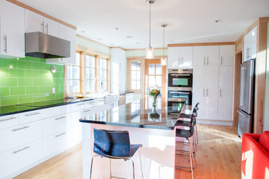 Example of a mid-sized danish l-shaped light wood floor open concept kitchen design in Minneapolis with a farmhouse sink, flat-panel cabinets, white cabinets, quartz countertops, green backsplash, cement tile backsplash, stainless steel appliances and an island