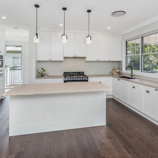 Неиссякаемый источник вдохновения для домашнего уюта: кухня среднего размера в классическом стиле с кладовкой, двойной раковиной, фасадами в стиле шейкер, белыми фасадами, столешницей из кварцевого композита, бежевым фартуком, фартуком из керамической плитки, черной техникой, островом, коричневым полом и розовой столешницей