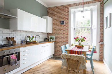 Photo of an eclectic single-wall eat-in kitchen in Edinburgh with a drop-in sink, flat-panel cabinets, white cabinets, white splashback, subway tile splashback, stainless steel appliances, medium hardwood floors, no island, brown floor, wood benchtops and brown benchtop.