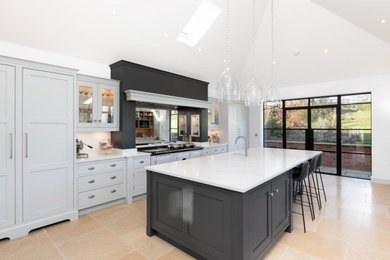 Expansive modern galley open plan kitchen in Hertfordshire with an integrated sink, shaker cabinets, white cabinets, engineered stone countertops, metallic splashback, mirror splashback, white appliances, porcelain flooring, an island, beige floors and white worktops.