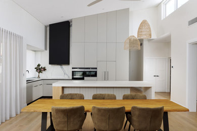 Photo of a large scandinavian l-shaped kitchen/diner in Perth with a double-bowl sink, flat-panel cabinets, grey cabinets, engineered stone countertops, white splashback, porcelain splashback, black appliances, light hardwood flooring, an island, beige floors, white worktops and a vaulted ceiling.
