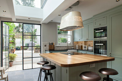 Mid-sized contemporary l-shaped kitchen in London with a farmhouse sink, shaker cabinets, green cabinets, wood benchtops, white splashback, subway tile splashback, black appliances, ceramic floors and with island.