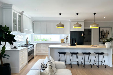 This is an example of a large modern l-shaped open plan kitchen in Sydney with shaker cabinets, white cabinets, quartz benchtops, with island, white benchtop, a farmhouse sink, white splashback, ceramic splashback, black appliances, light hardwood floors and brown floor.