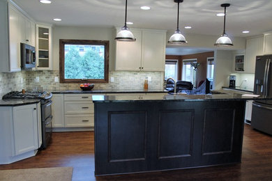 Example of a large classic u-shaped dark wood floor and brown floor enclosed kitchen design in Milwaukee with recessed-panel cabinets, white cabinets, granite countertops, gray backsplash, stone tile backsplash, stainless steel appliances and an island