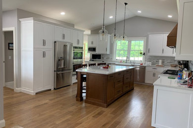 Mid-sized farmhouse l-shaped light wood floor and brown floor enclosed kitchen photo in Other with recessed-panel cabinets, white cabinets, gray backsplash, glass tile backsplash, stainless steel appliances, an island, white countertops, a farmhouse sink and quartzite countertops