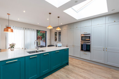 Photo of a large contemporary grey and teal l-shaped open plan kitchen in London with a submerged sink, shaker cabinets, green cabinets, quartz worktops, integrated appliances, vinyl flooring, an island and white worktops.