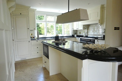 Photo of a traditional kitchen/diner in West Midlands with a double-bowl sink, raised-panel cabinets, white cabinets, granite worktops, white splashback, ceramic splashback, black appliances, limestone flooring and an island.