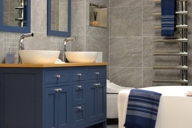 This is an example of a mid-sized traditional kids bathroom in Other with beaded inset cabinets, blue cabinets, a freestanding tub, a wall-mount toilet, gray tile, porcelain tile, grey walls, porcelain floors, a vessel sink, wood benchtops, grey floor and brown benchtops.