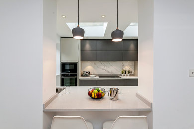 Photo of a contemporary galley open plan kitchen in London with a drop-in sink, flat-panel cabinets, grey cabinets, quartzite benchtops, metallic splashback, stone slab splashback, black appliances and a peninsula.