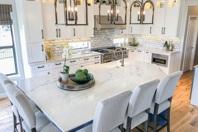 Example of a mid-sized beach style light wood floor and brown floor eat-in kitchen design in Houston with a farmhouse sink, recessed-panel cabinets, white cabinets, quartzite countertops, gray backsplash, ceramic backsplash, stainless steel appliances, an island and white countertops