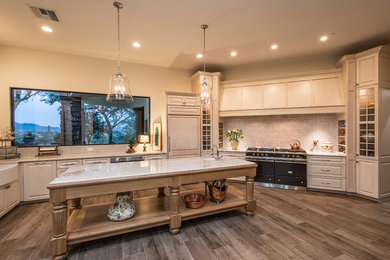 Example of a large farmhouse u-shaped travertine floor and brown floor open concept kitchen design in Phoenix with raised-panel cabinets, an island, beige cabinets, marble countertops, white countertops, a farmhouse sink and paneled appliances