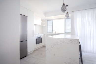 Small beach style u-shaped eat-in kitchen in Sunshine Coast with a single-bowl sink, white cabinets, solid surface benchtops, white splashback, glass sheet splashback, stainless steel appliances, laminate floors, with island, grey floor and white benchtop.