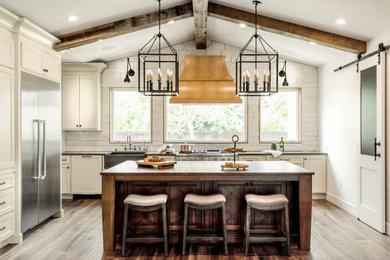 Inspiration for a mediterranean l-shaped kitchen in Austin with a farmhouse sink, shaker cabinets, white cabinets, white splashback, stainless steel appliances, medium hardwood floors, with island, brown floor, black benchtop, exposed beam and vaulted.