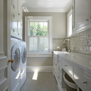 ニューヨークの中サイズのトラディショナルスタイルのおしゃれな洗濯室 (白いキャビネット、エプロンフロントシンク、人工大理石カウンター、セラミックタイルの床、左右配置の洗濯機・乾燥機、グレーの床、白いキッチンカウンター、インセット扉のキャビネット、グレーの壁) の写真