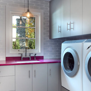 グランドラピッズの大きいコンテンポラリースタイルのおしゃれな家事室 (左右配置の洗濯機・乾燥機、アンダーカウンターシンク、フラットパネル扉のキャビネット、グレーのキャビネット、ラミネートカウンター、グレーの壁、セラミックタイルの床、グレーの床、ピンクのキッチンカウンター) の写真