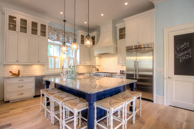 Example of a huge beach style l-shaped light wood floor open concept kitchen design in Miami with a farmhouse sink, shaker cabinets, white cabinets, granite countertops, white backsplash, ceramic backsplash, stainless steel appliances and an island